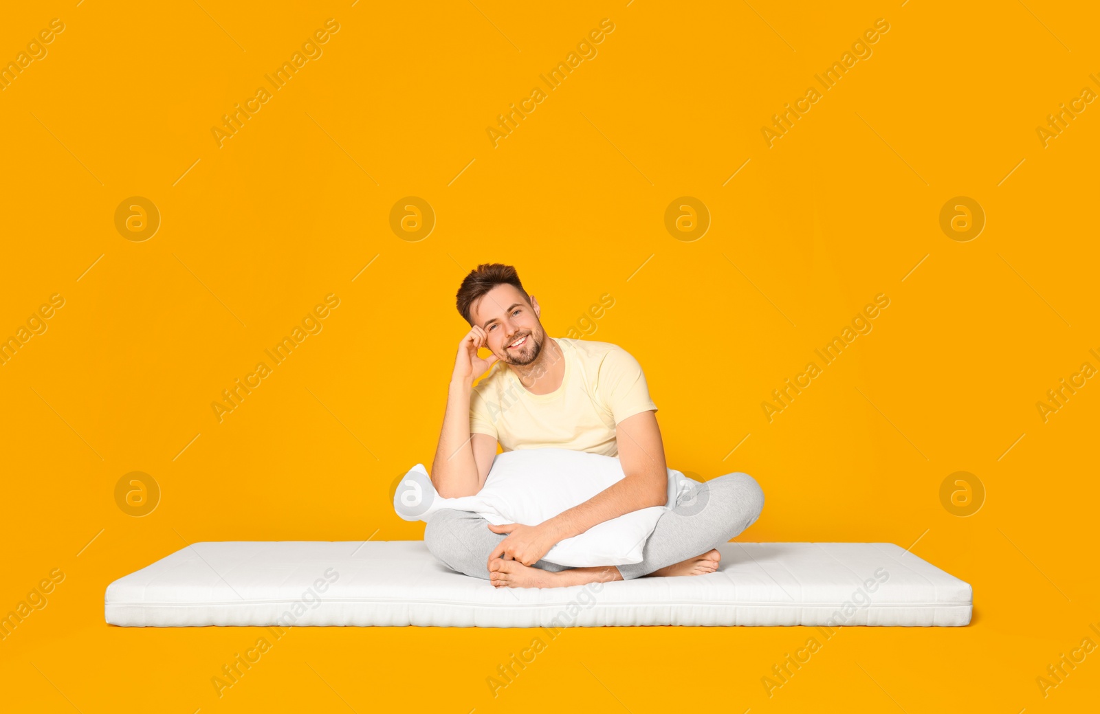 Photo of Smiling man with pillow sitting on soft mattress against orange background