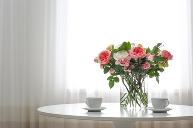 Vase with beautiful flower bouquet and cups of coffee on table in room. Space for text