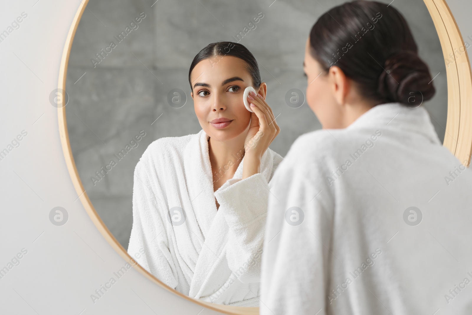 Photo of Beautiful woman removing makeup with cotton pad near mirror indoors