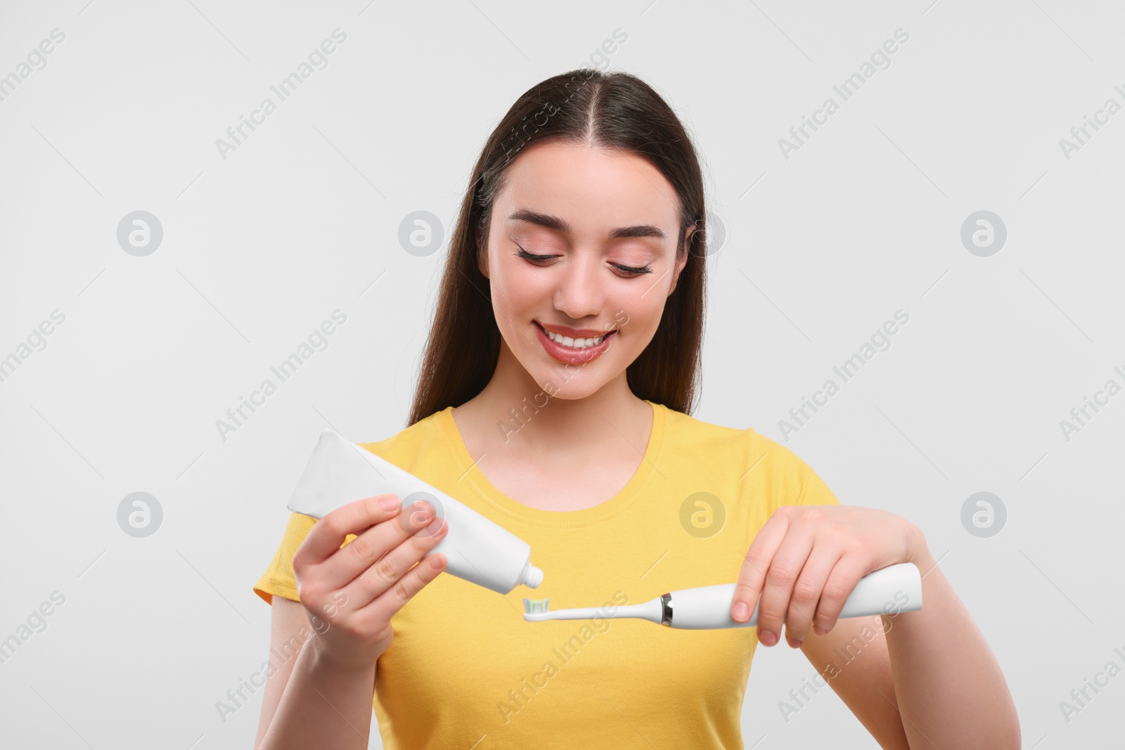 Photo of Happy young woman squeezing toothpaste from tube onto electric toothbrush on white background