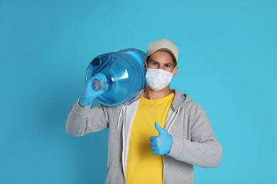 Photo of Courier in medical mask with bottle for water cooler showing thumb up on light blue background. Delivery during coronavirus quarantine
