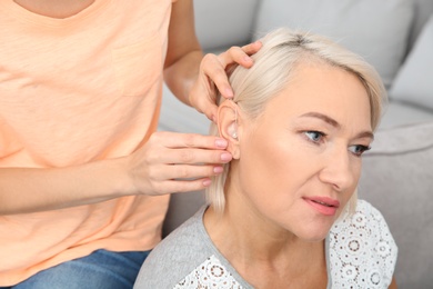 Young woman putting hearing aid in mother's ear indoors