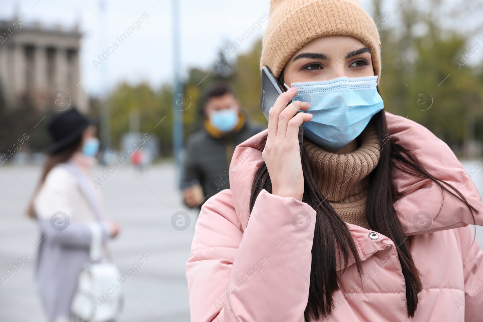 Photo of Young woman in medical face mask talking on phone while walking outdoors. Personal protection during COVID-19 pandemic