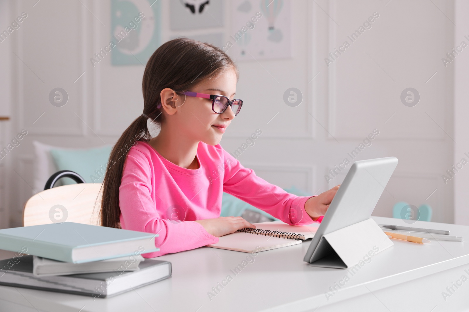 Photo of Little girl doing homework with tablet at table in room