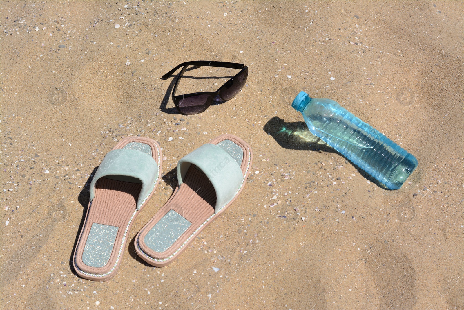 Photo of Stylish sunglasses, slippers and bottle of water on sand. Beach accessories