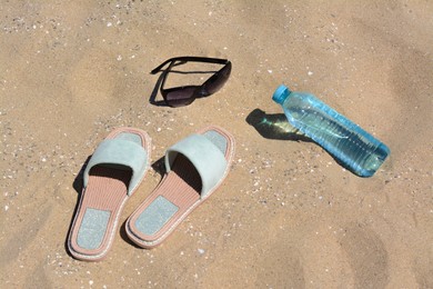 Photo of Stylish sunglasses, slippers and bottle of water on sand. Beach accessories