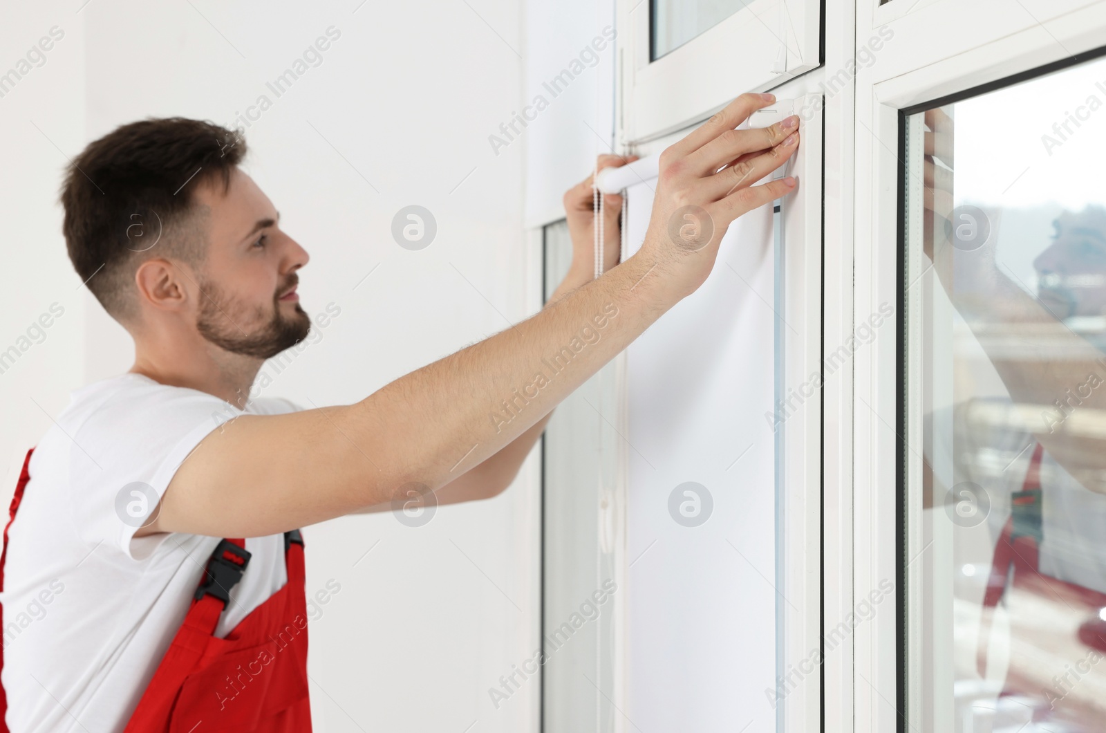 Photo of Worker in uniform installing roller window blind indoors, focus on hands