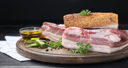 Photo of Pieces of raw pork belly, chili pepper, peppercorns, oil and parsley on black wooden table, closeup