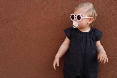 Photo of Cute little girl wearing stylish clothes with sunglasses and pacifier near brown wall. Space for text
