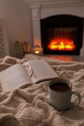 Photo of Cup of hot tea, eyeglasses and book near fireplace at home. Cozy atmosphere