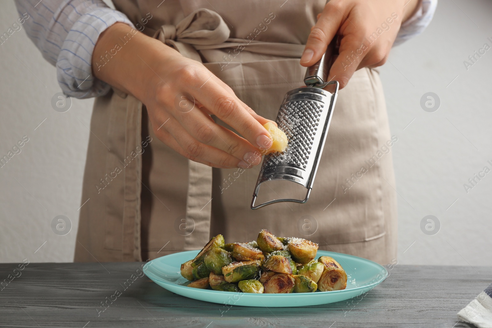 Photo of Woman grating cheese on roasted brussels sprouts at grey wooden table, closeup