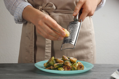 Woman grating cheese on roasted brussels sprouts at grey wooden table, closeup