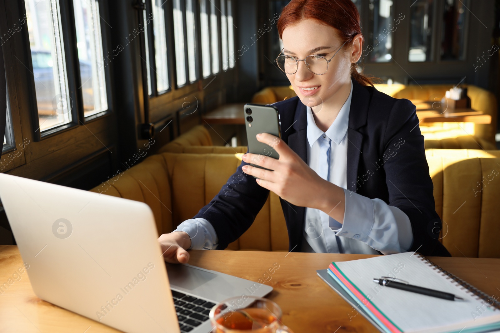 Photo of Young female student with laptop using smartphone while studying at table in cafe