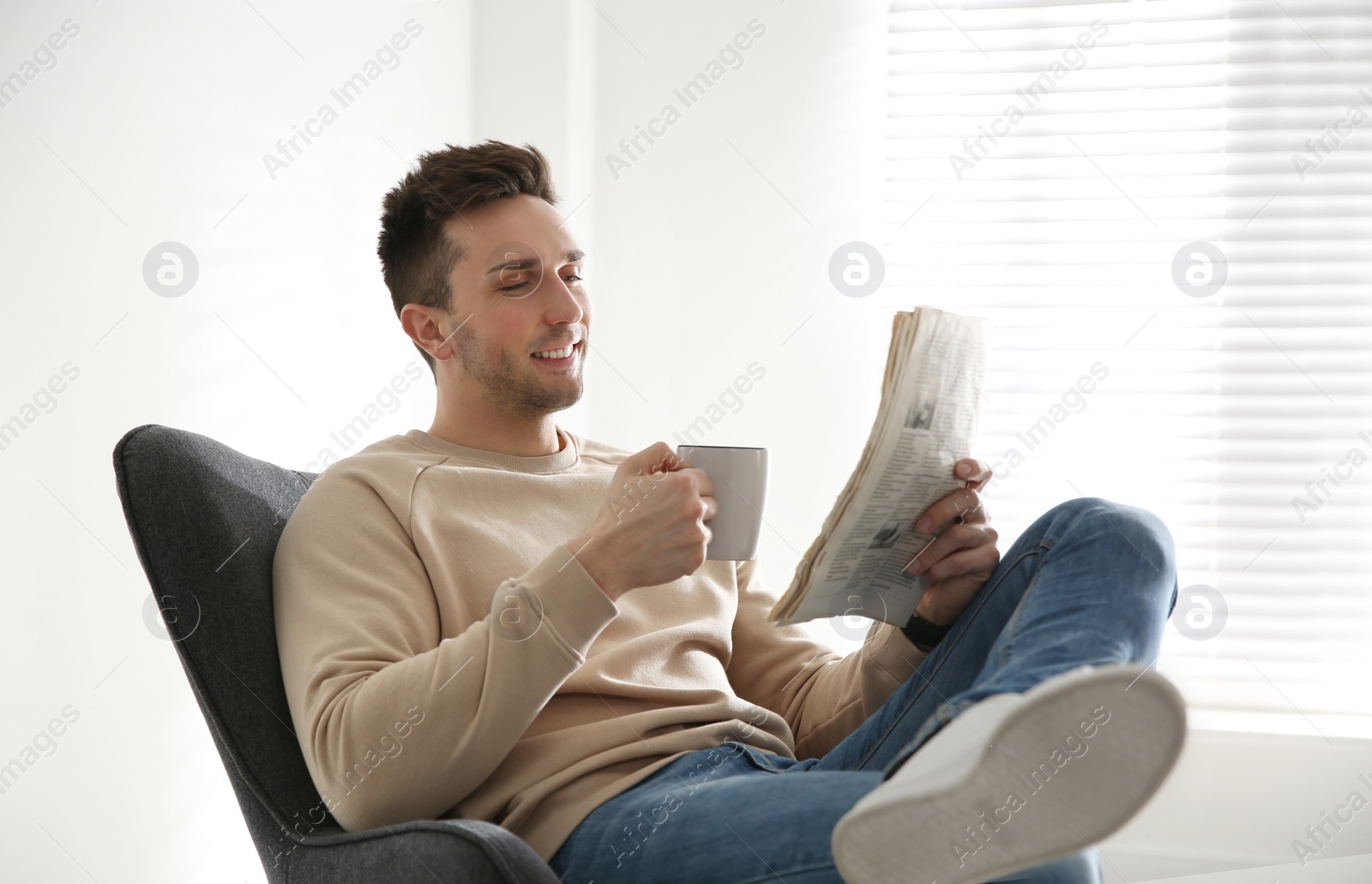 Photo of Young man with cup of drink and newspaper relaxing in armchair near window at home