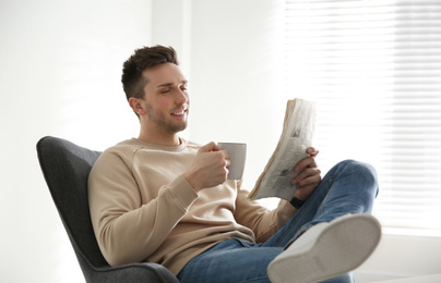 Young man with cup of drink and newspaper relaxing in armchair near window at home