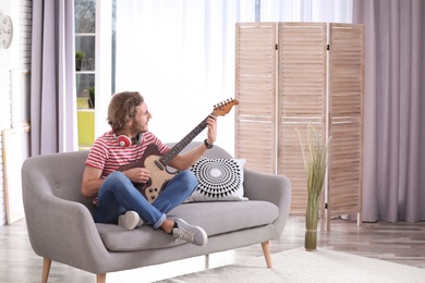Young man with headphones playing electric guitar in living room. Space for text