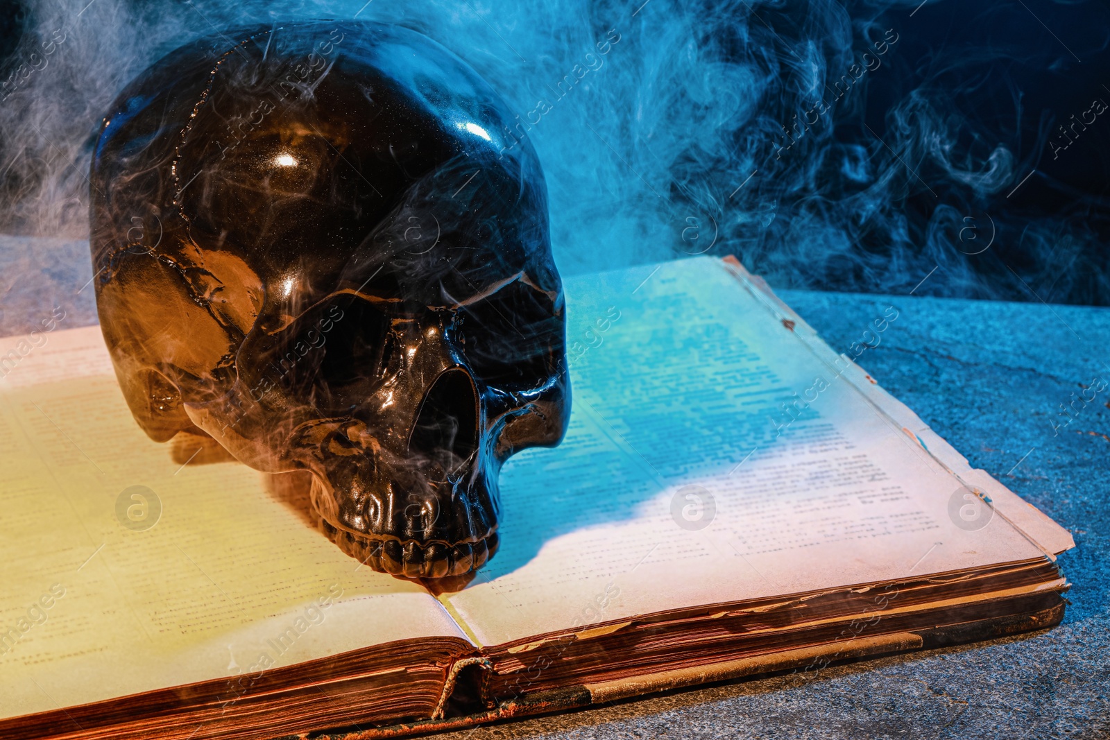 Photo of Black human skull, old book and smoke on stone surface against color background