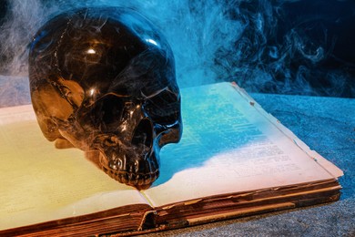 Black human skull, old book and smoke on stone surface against color background