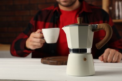 Man with cup of drink at white table indoors, focus on coffee moka pot