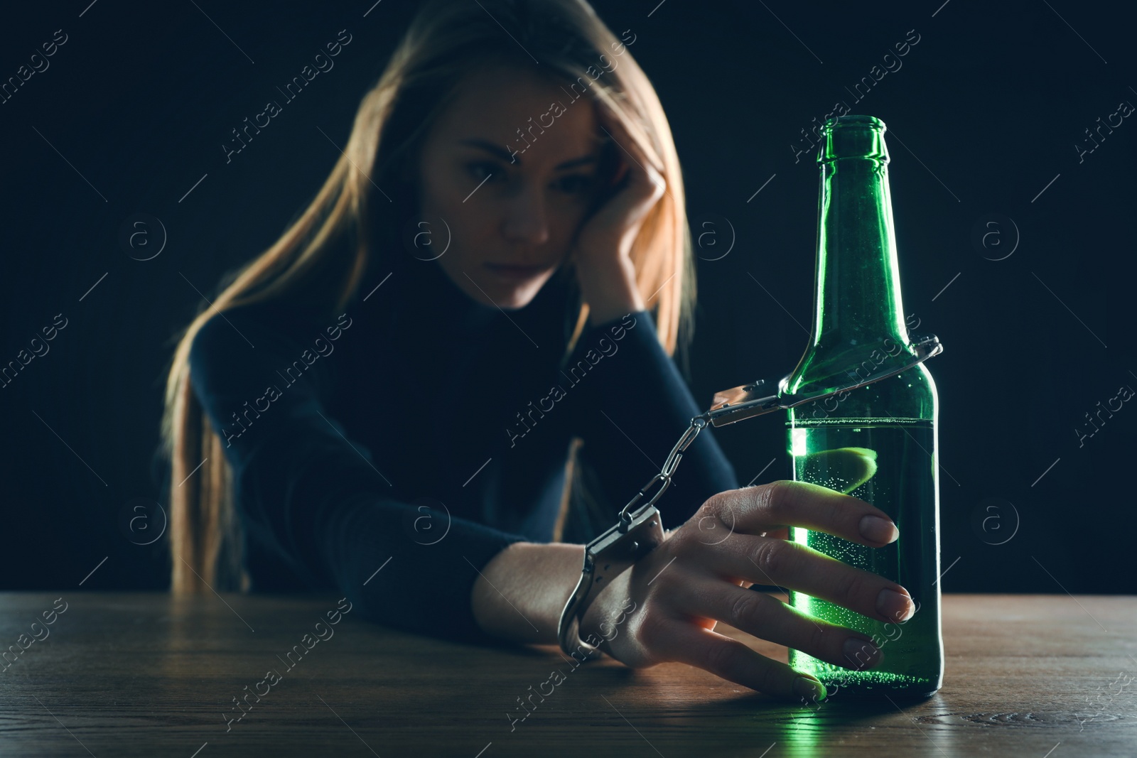 Photo of Alcohol addiction. Woman handcuffed to bottle of beer at wooden table against black background, focus on hand