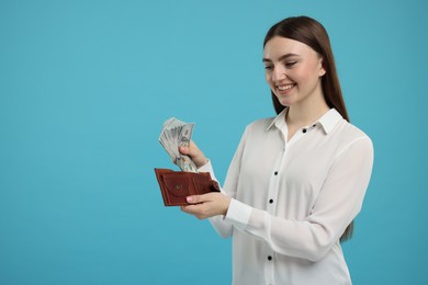 Happy woman putting money into wallet on light blue background, space for text