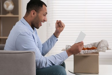 Emotional man with greeting card near parcel at home. Internet shopping