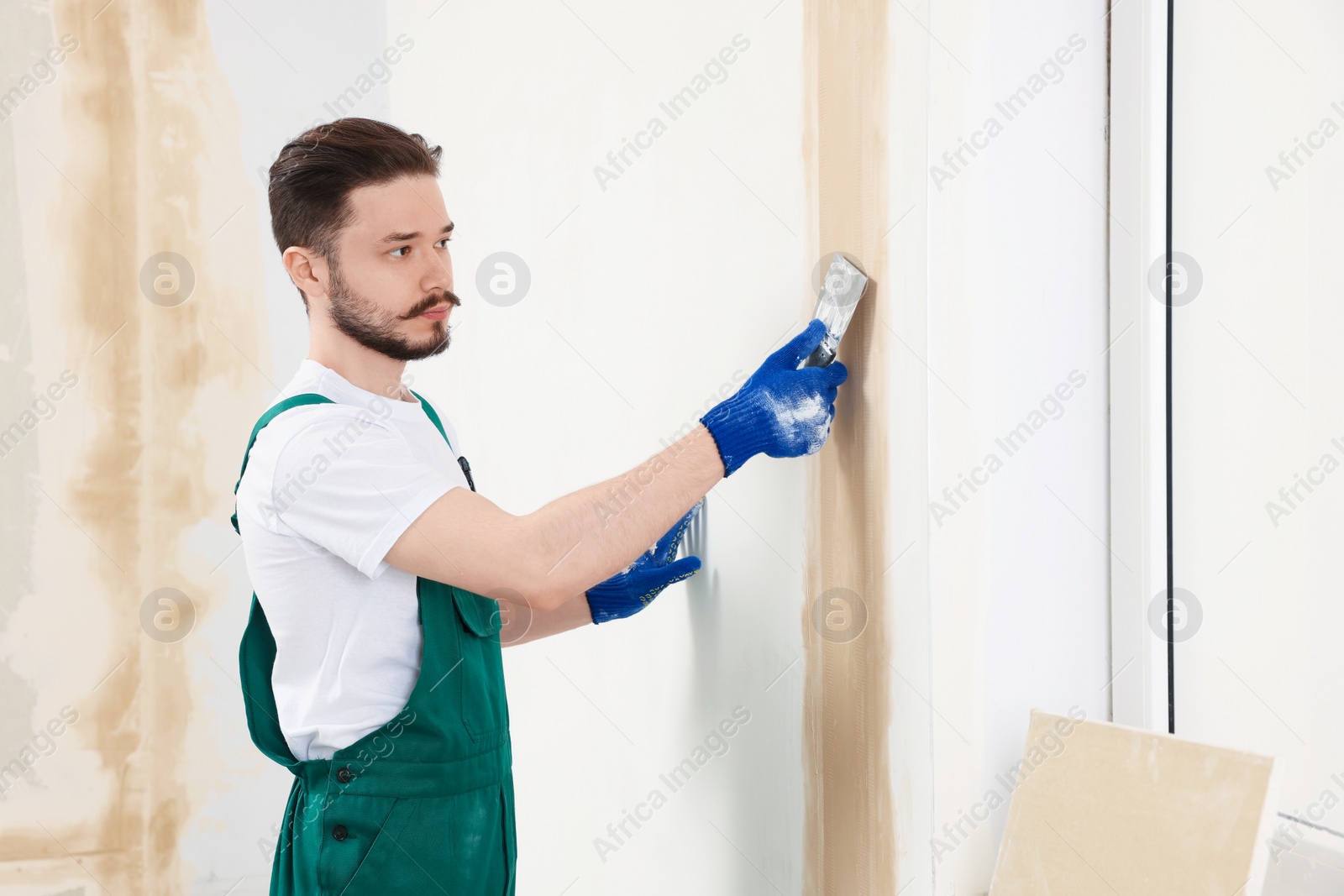 Photo of Worker in uniform plastering wall with putty knife indoors