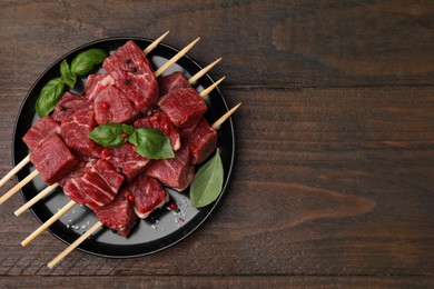 Photo of Skewers with cut fresh beef meat, basil leaves and spices on wooden table, top view. Space for text