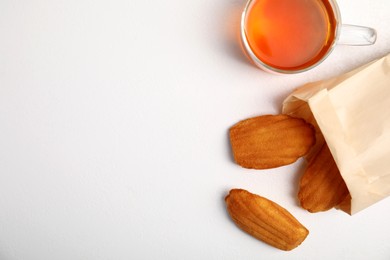 Photo of Paper bag with delicious madeleine cakes and cup of tea on white background, flat lay. Space for text