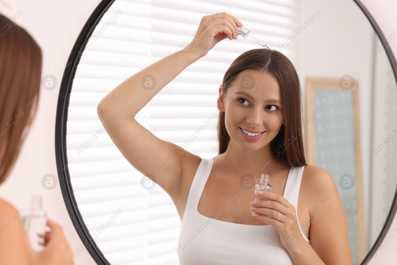 Photo of Beautiful woman applying serum onto hair near mirror indoors