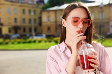 Photo of Young woman in sunglasses with plastic cup of fresh juice on city street, space for text