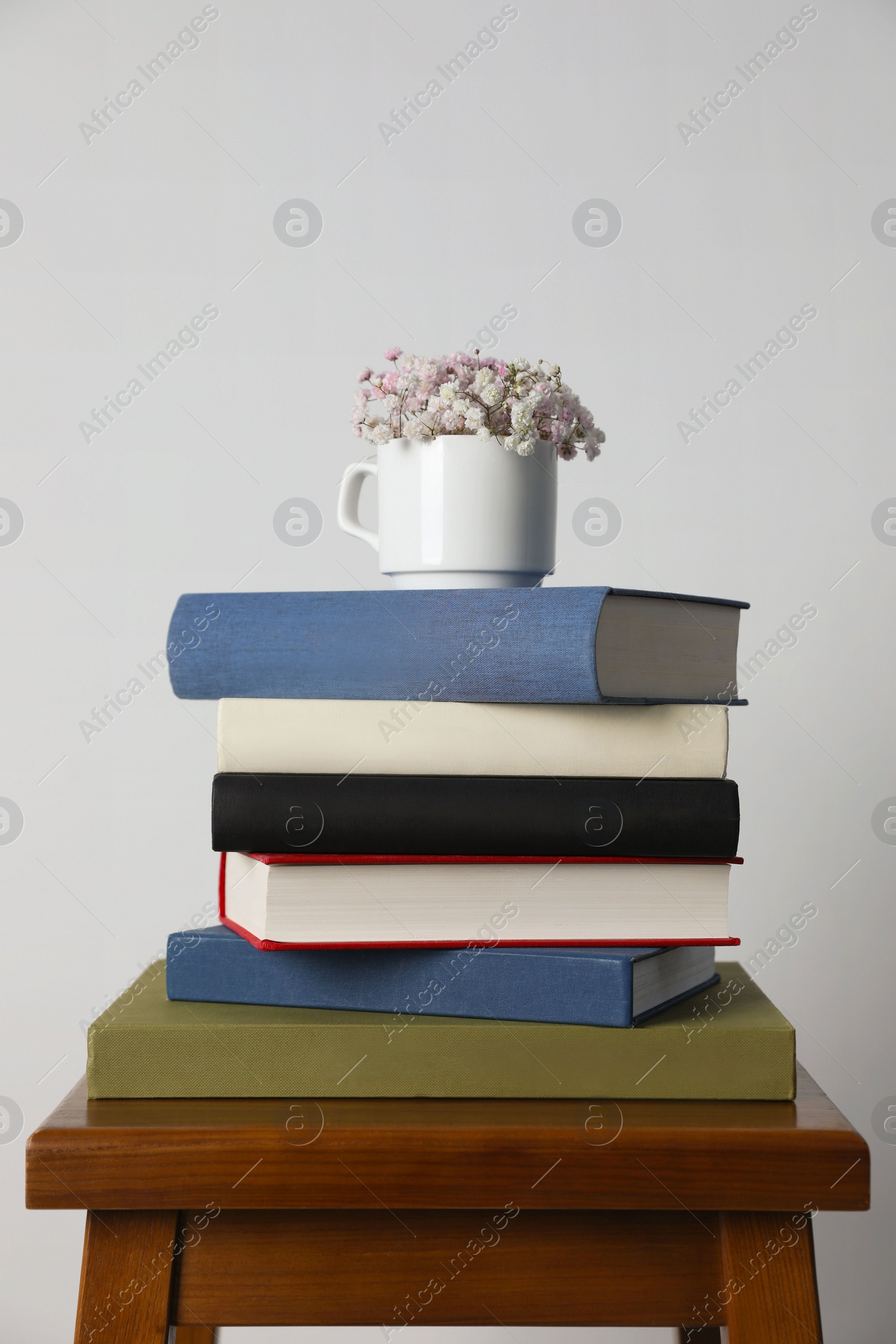 Photo of Hardcover books and cup with flowers on wooden stool near white wall