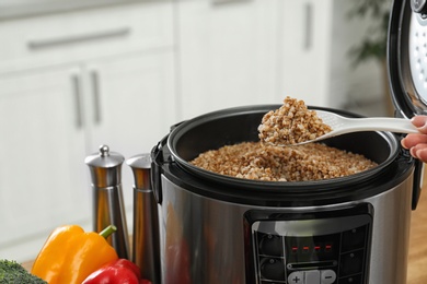 Photo of Woman holding spoon with buckwheat over multi cooker in kitchen, closeup view