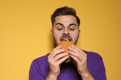 Handsome man eating tasty burger on color background
