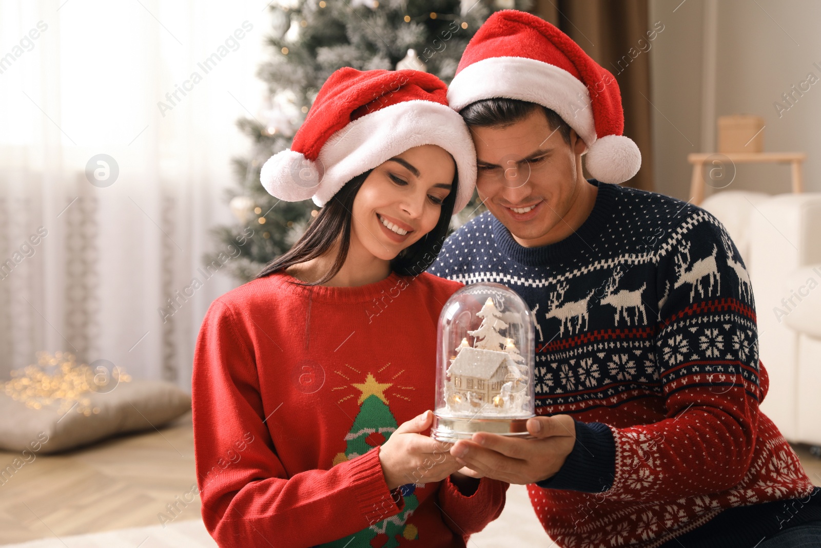 Photo of Couple in Santa hats holding snow globe at home