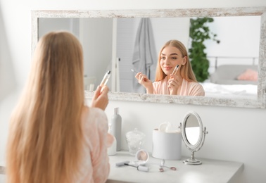 Young woman applying oil onto her eyelashes near mirror indoors