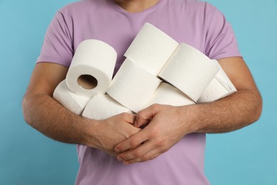 Man with heap of toilet paper rolls on light blue background, closeup