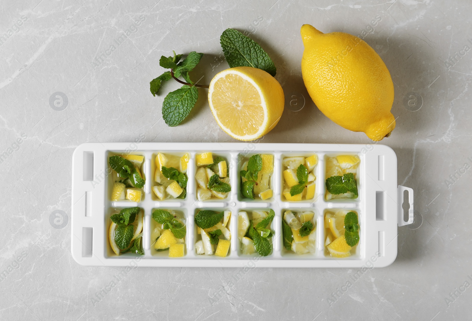 Photo of Ice cube tray with mint, lemon and water on grey background, top view