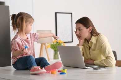 Photo of Woman working remotely at home. Little girl bothering her mother. Child sitting on desk