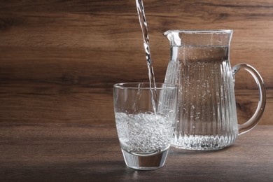Photo of Pouring soda water into glass near jug on wooden table. Space for text