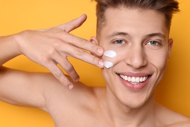 Photo of Handsome man applying moisturizing cream onto his face on orange background, closeup