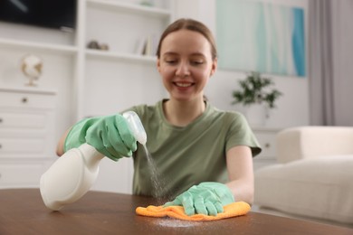 Photo of Woman with spray bottle and microfiber cloth cleaning wooden table in room, selective focus