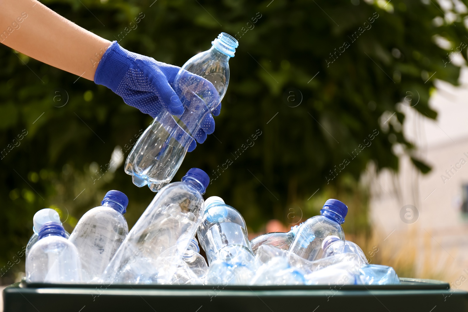 Photo of Woman in gloves putting used plastic bottle into trash bin outdoors, closeup. Recycling problem