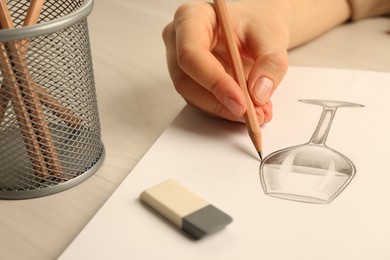 Woman drawing glass of wine with graphite pencil at light wooden table, closeup