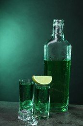 Photo of Absinthe in shot glasses with lime and ice cubes on gray textured table against green background. Alcoholic drink