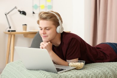 Photo of Teenage boy with headphones and popcorn using laptop on bed at home
