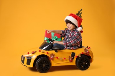 Photo of Cute little boy in Santa hat with Christmas tree and gift box driving children's electric toy car on yellow background