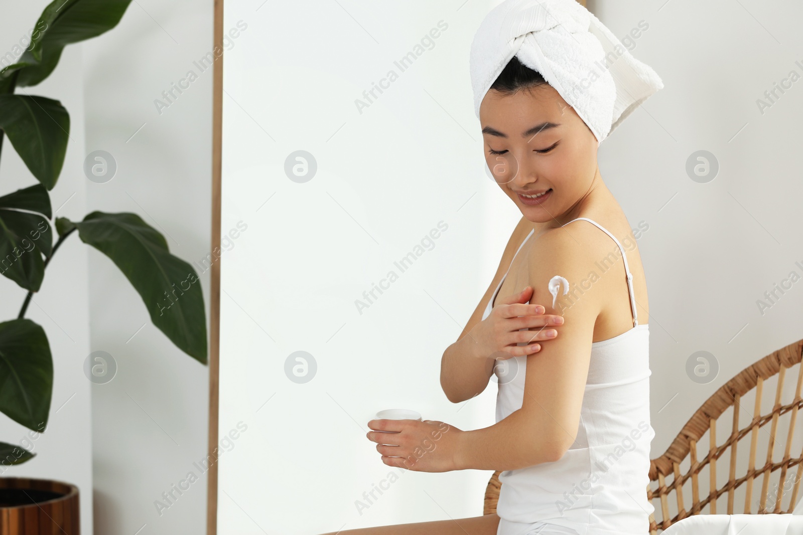 Photo of Beautiful young Asian woman applying body cream on shoulder in room, space for text