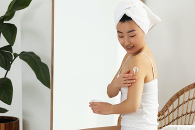 Beautiful young Asian woman applying body cream on shoulder in room, space for text
