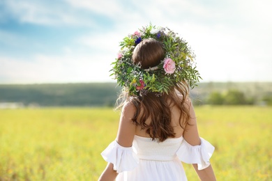 Young woman wearing wreath made of beautiful flowers in field on sunny day, back view